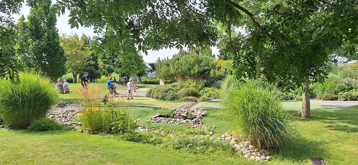 Rasen und Steinflächen mit Personen die auf einer Bank sitzen oder auch welche die Abenteuergolf spielen mit Büschen und Bäumen. Im Hintergrund sind Gebäude vom Erlebnsipark und dem Ocean wave zu sehen.