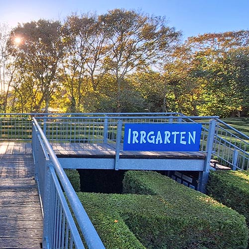 Grüne Hecken im Irrgarten mit Brücke aus Holz und Metall mit Personen drauf und die Sonne scheint.